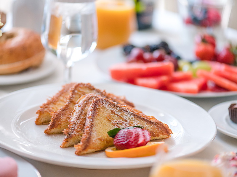 Plate of French toast garnished with strawberries and orange slices, surrounded by fresh fruit and pastries in a breakfast setting.